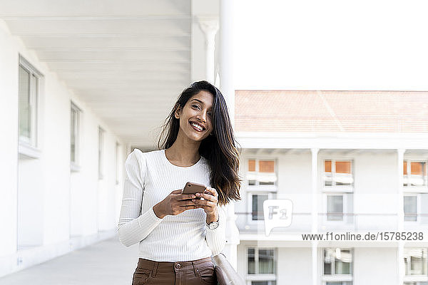 Lächelnde junge Frau mit Smartphone in der Hand auf dem Balkon