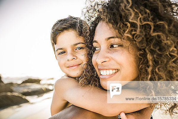 Porträt einer glücklichen Mutter  die ihren Sohn huckepack am Strand trägt