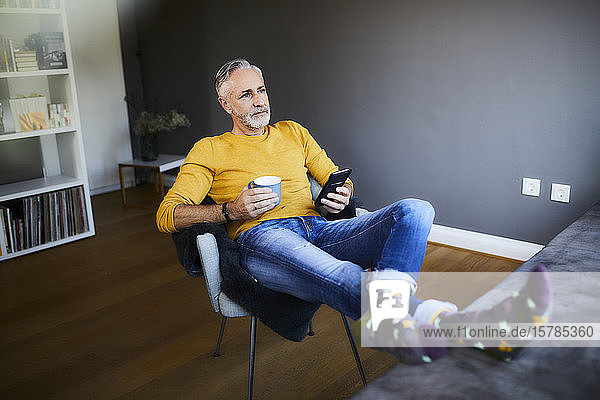 Relaxed mature man at home with cell phone and cup of coffee