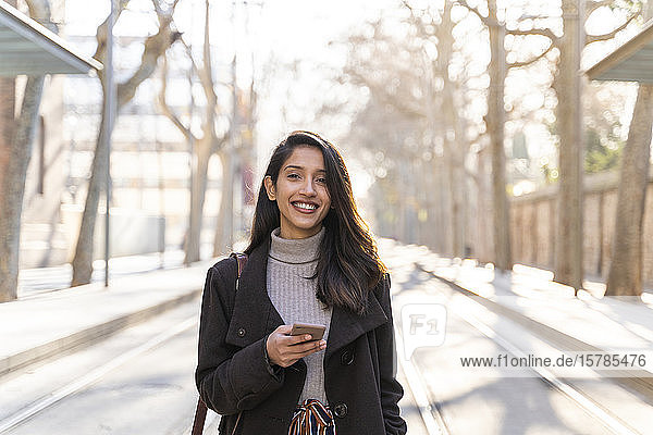 Porträt einer lächelnden jungen Frau mit Smartphone auf einer Straßenbahnlinie