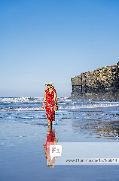 Blonde Frau in rotem Kleid und Hut die am Strand spazieren geht Playa de Las Catedrales Spanien