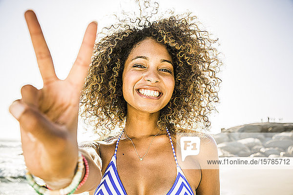 Porträt einer glücklichen jungen Frau beim Siegeszeichen am Strand