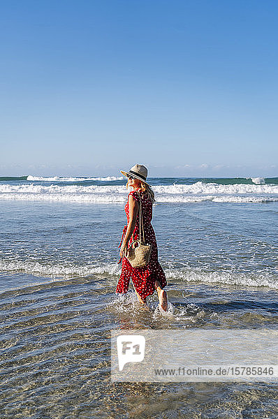 Blonde Frau in rotem Kleid und Hut  die am Strand spazieren geht  Playa de Las Catedrales  Spanien