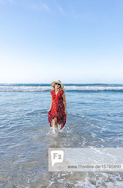 Blonde Frau mit rotem Kleid und Hut am Strand  die im Wasser läuft
