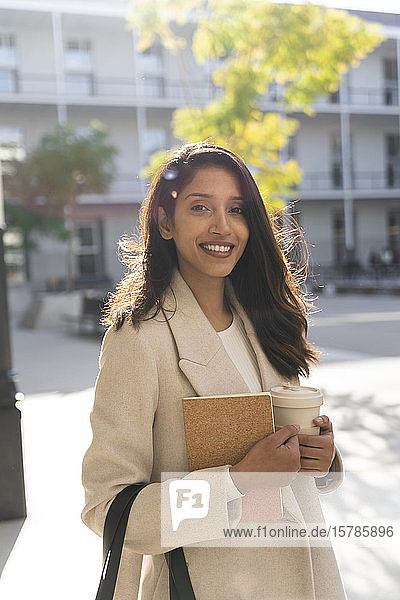 Portrait of smiling young woman with book and takeaway coffee in the city