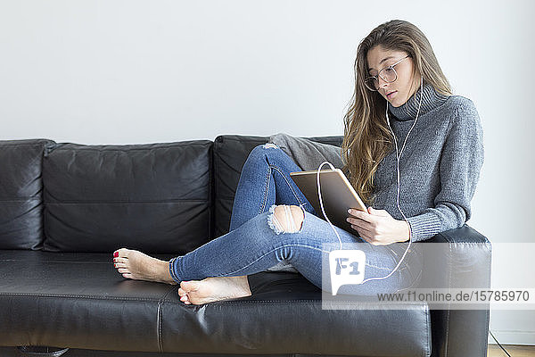 Junge Frau sitzt auf schwarzer Ledercouch mit Kopfhörern und digitalem Tablet