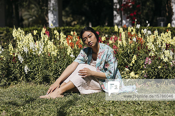 Schöne junge Frau sitzt im Frühling im Gras  umgeben von Blumen in einem öffentlichen Garten