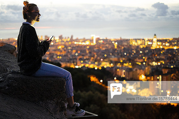 Young woman sitting on railing above the city using cell phone  Barcelona  Spain