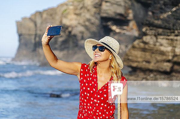 Blonde Frau mit rotem Kleid und Hut  die ein Smartphone benutzt und am Strand ein Selfie macht