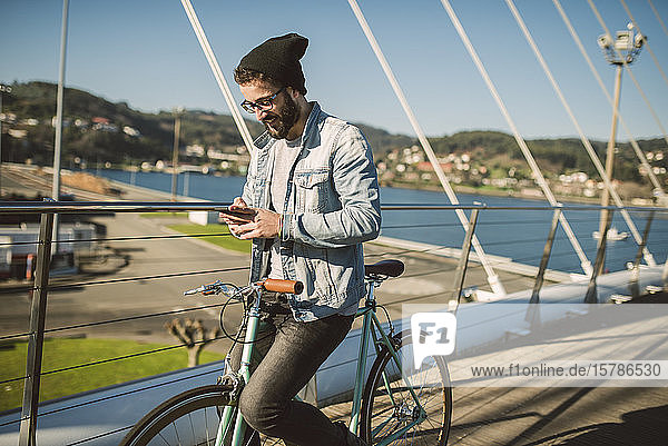 Junger Mann der mit seinem Fixie-Rad durch die Stadt pendelt und ein Smartphone benutzt