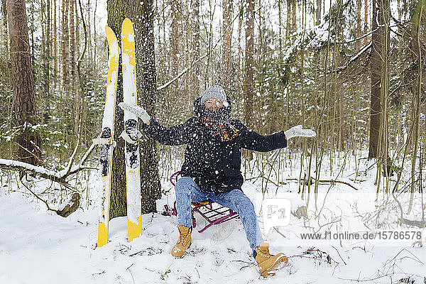 Woman with skis sitting on sledge in winter forest throwing snow into the air