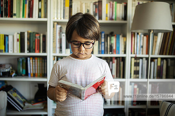 Portrait of serious boy with glasses standing in front of book shelves reading a comic