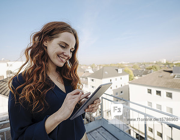 Lächelnde rothaarige Frau mit Tablette auf der Dachterrasse