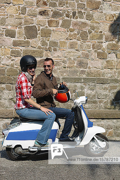 Laughing couple on motor scooter in front of stone wall  Italy
