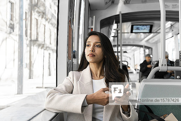 Junge Frau mit Smartphone  die in einer Straßenbahn aus dem Fenster schaut