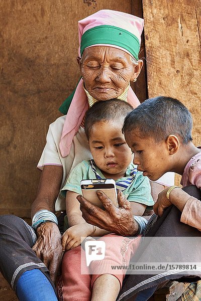 Kayan Lahwi woman with brass neck coils and traditional clothing looking at cellphone with her grandchildren. The Long Neck Kayan (also called Padaung in Burmese) are a sub-group of the Karen ethnic from Burma. Women wear spiral coils around their neck and lower legs. They are also nicknamed 'giraffe womenâ. œ. The neck itself is not lengthened  the appearance of a stretched neck is created under the pressure of the collar  the ribs lower and the shoulders and clavicles collapse. Elderly people generally look after the children while the parents are working in the farm. Pan Pet Region  Kayah State  Myanmar.
