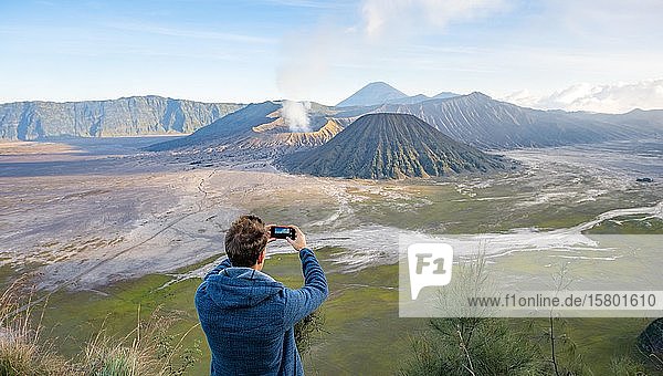 Young man takes photo with iPhone  volcanic landscape  view in Tengger Caldera  smoking volcano Gunung Bromo  in front Mt. Batok  in the back Mt. Kursi  Mt. Gunung Semeru  National Park Bromo-Tengger-Semeru  Java  Indonesia  Asia
