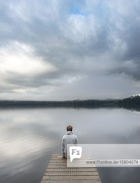Young man sitting on a jetty looking over a lake  foggy atmosphere  Lake Mapourika  West Coast  South Island  New Zealand  Oceania