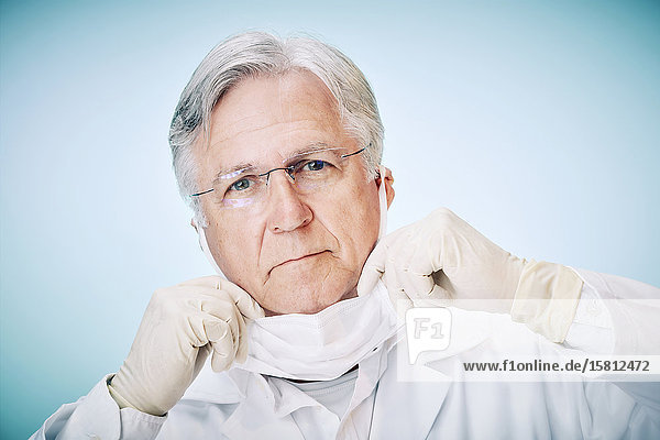 Grauhaariger Arzt mit Schutzhandschuhen und Schutzbrille zieht Mundschutz hoch