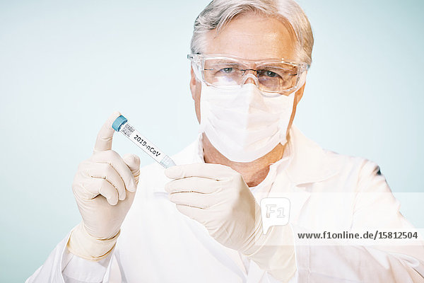 Grauhaariger Laborant mit Schutzbrille und Mundschutz blickt auf Reagenzglas mit Cornoravirus Probe