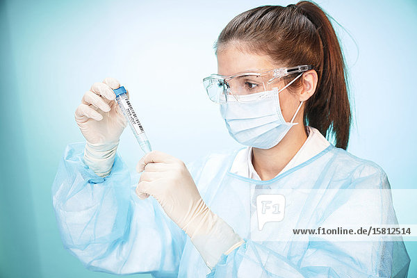 Laborantin mit Schutzbrille und Mundschutz blickt auf Reagenzglas mit Cornavirus Probe