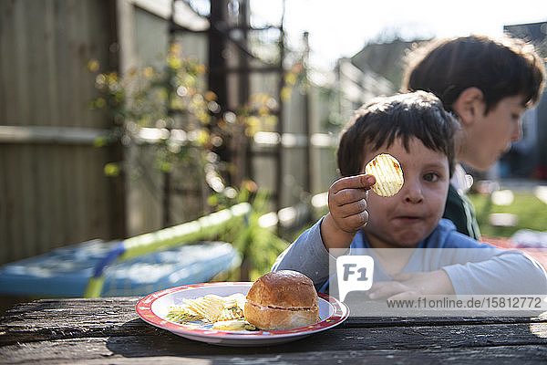 Porträt von zwei Jungen  die an einem Tisch in einem Garten sitzen und einen Snack mit Burger und Chips essen.