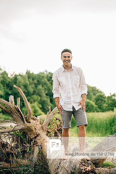 Junger Mann in Shorts steht auf umgestürztem Baum am Flussufer und lächelt in die Kamera.