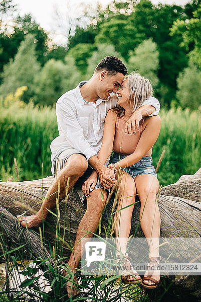 Junges Paar sitzt auf einem umgefallenen Baum am Flussufer  lächelt und umarmt sich.