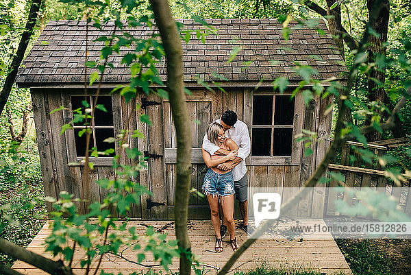 Junges Paar steht vor einer Holzhütte  umarmt sich und lächelt.