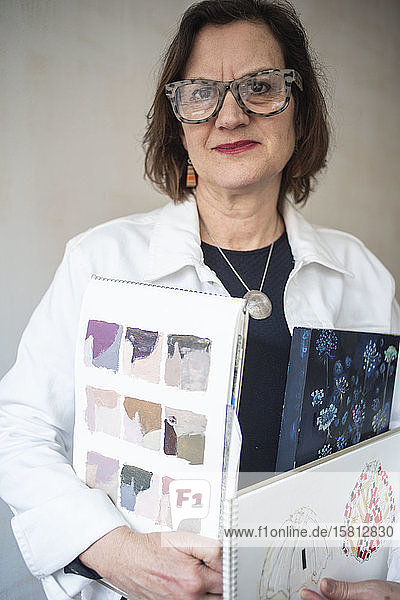 Portrait of brunette woman wearing glasses and white denim jacket  holding artist's sketch books.