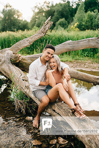 Junges Paar sitzt auf einem umgefallenen Baum am Flussufer  lächelt und umarmt sich.