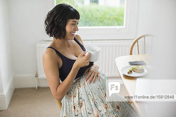 Glückliche schwangere Frau trinkt Tee am Laptop