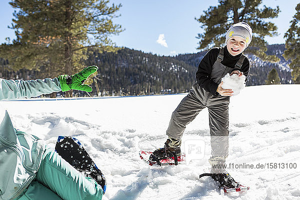 Ein sechsjähriger Junge mit Schneeschuhen  der einen großen Schneeball hält.