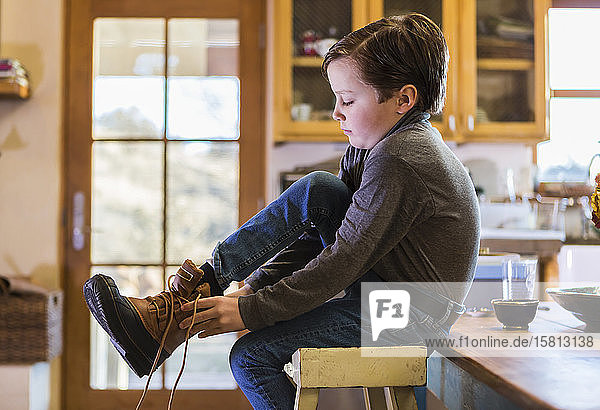 Ein sechsjähriger Junge  der seine Stiefel anzieht und auf einem hohen Schemel sitzt.