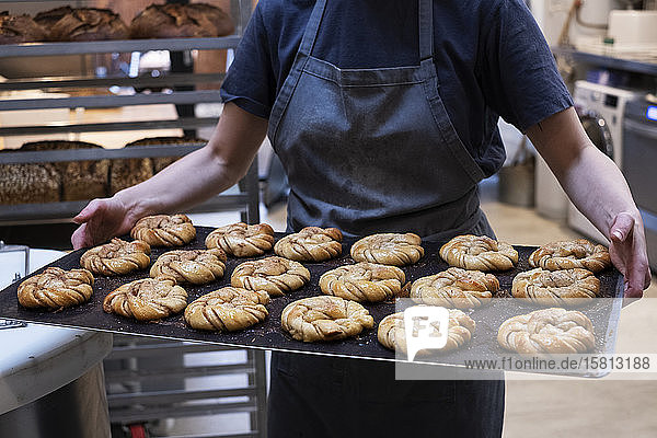 Nahaufnahme einer Person  die in einer handwerklichen Bäckerei ein Tablett mit frisch gebackenen Zimtbrötchen hält.