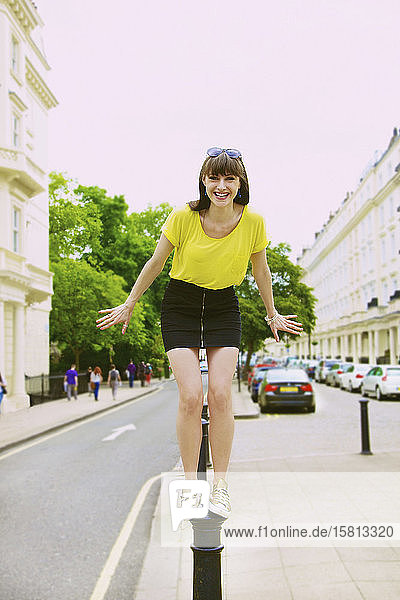 Porträt einer fröhlichen  verspielten jungen Frau  die auf einem Straßenpfosten steht