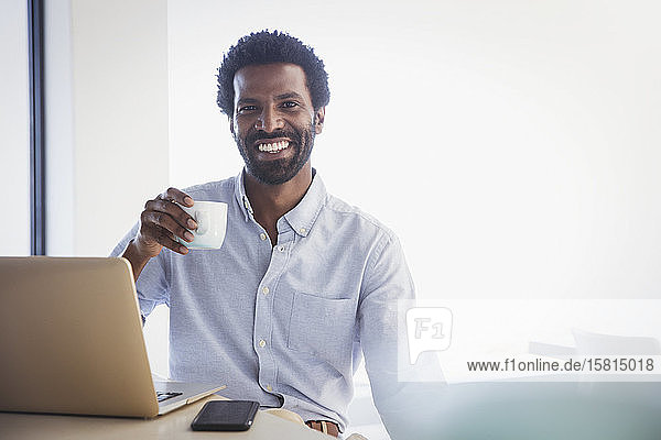 Porträt eines lächelnden  selbstbewussten Geschäftsmannes  der am Laptop Kaffee trinkt