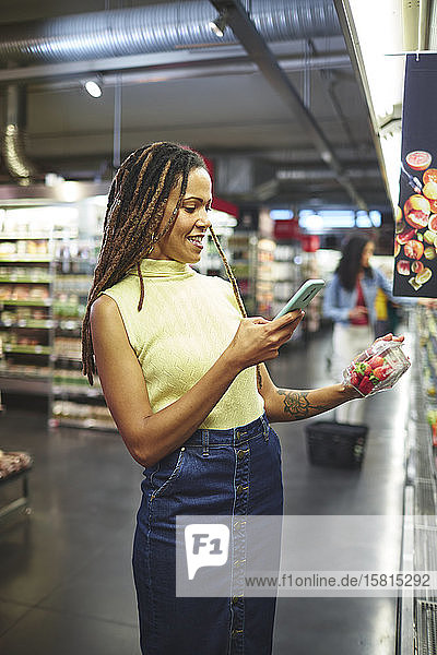 Lächelnde Frau mit Smartphone beim Lebensmitteleinkauf im Supermarkt