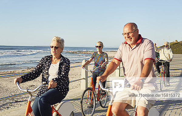 Active senior tourist friends bike riding on sunny boardwalk along ocean
