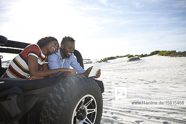 Junges Paar  das ein digitales Tablet im Jeep am sonnigen Strand benutzt und einen Roadtrip genießt