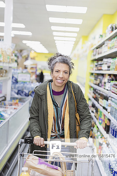 Porträt einer lächelnden  selbstbewussten Frau  die einen Einkaufswagen in einem Supermarktgang schiebt