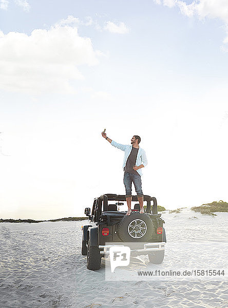 Junger Mann mit Fotohandy,  der ein Selfie auf einem Jeep-Reifen am Strand macht
