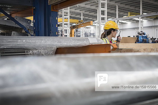 Female worker at laptop talking on walkie-talkie in factory