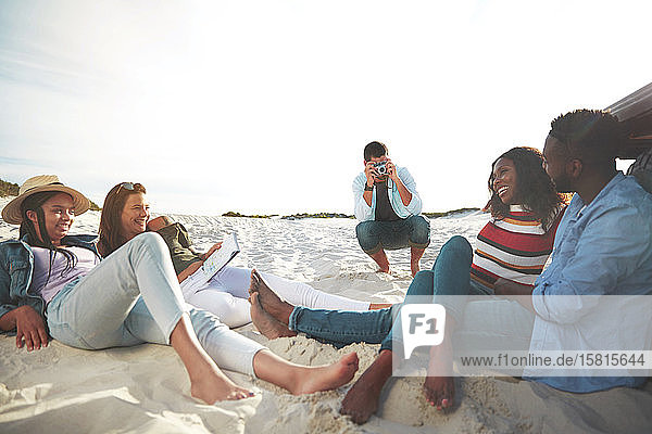 Junger Mann mit Digitalkamera fotografiert Freunde beim Entspannen am Strand