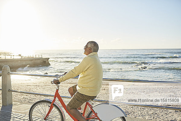 Aktiver älterer Mann  der auf der sonnigen Strandpromenade am Meer Fahrrad fährt