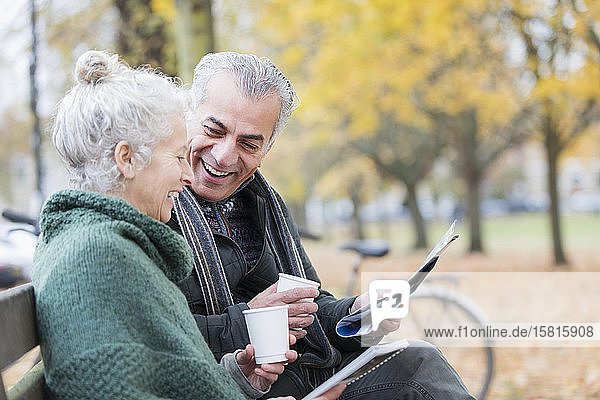Lächelndes älteres Paar liest Zeitung und trinkt Kaffee auf einer Bank im Herbstpark