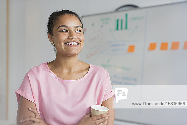 Porträt einer lächelnden  ehrgeizigen Geschäftsfrau  die am Whiteboard im Büro Kaffee trinkt