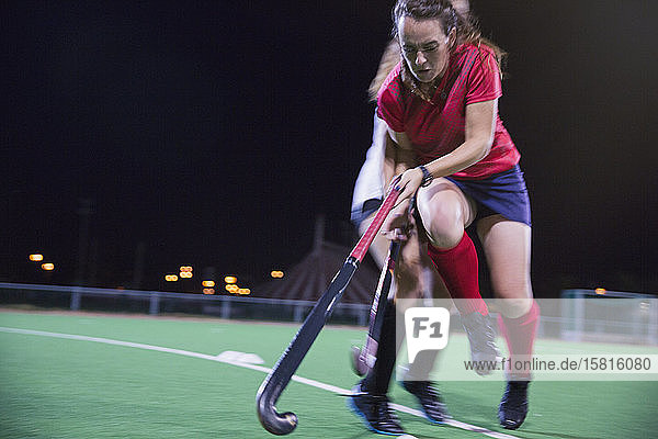 Determined young female field hockey player reaching with hockey stick  playing on field at night