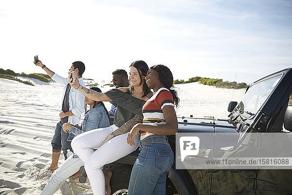 Young friends with camera phones taking selfie at jeep on sunny beach