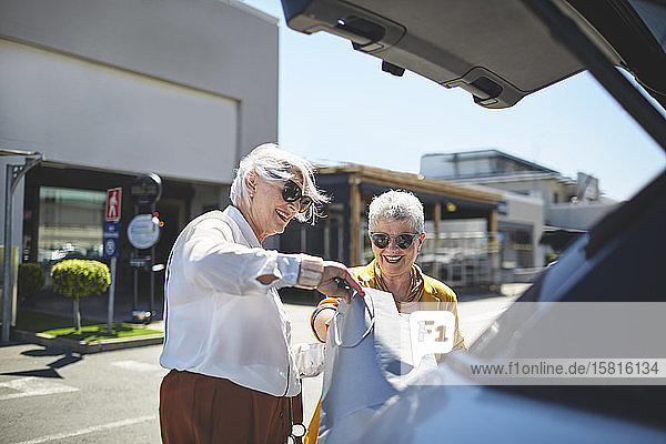 Ältere Freundinnen laden Einkaufstüten auf einem sonnigen Parkplatz in den Kofferraum eines Autos